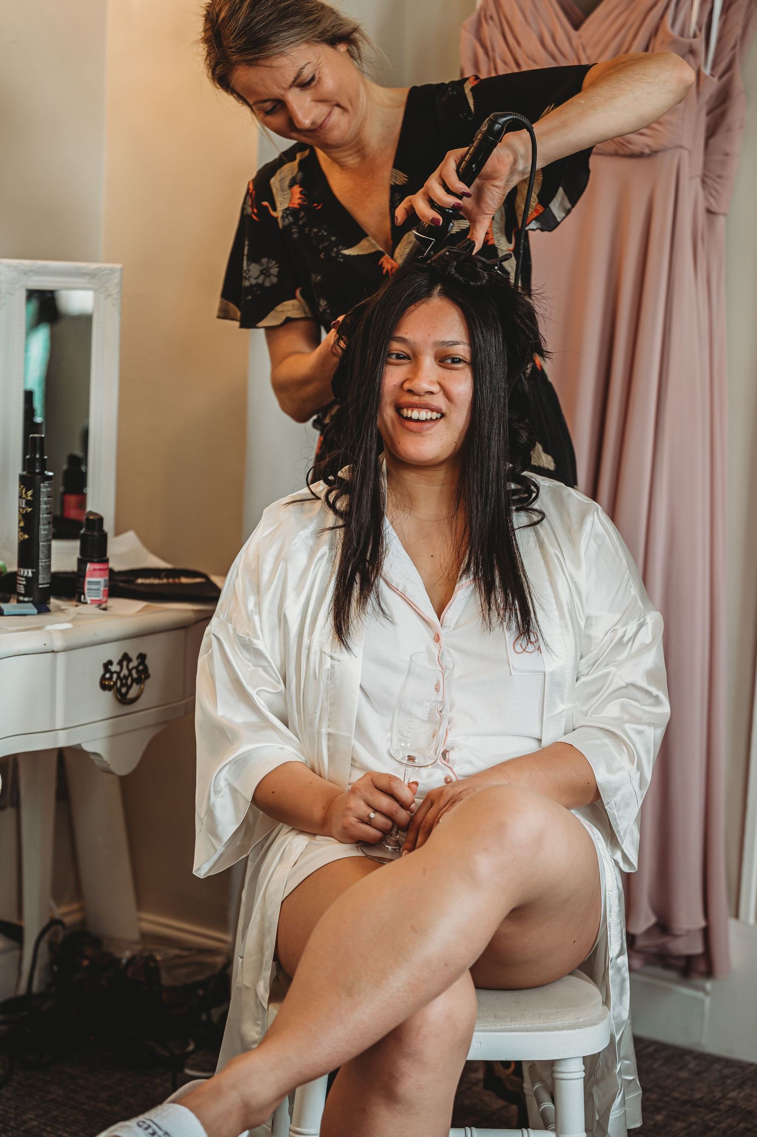 A hairstylist styles a client's hair in a bridal preparation setting with natural lighting and soft decor.