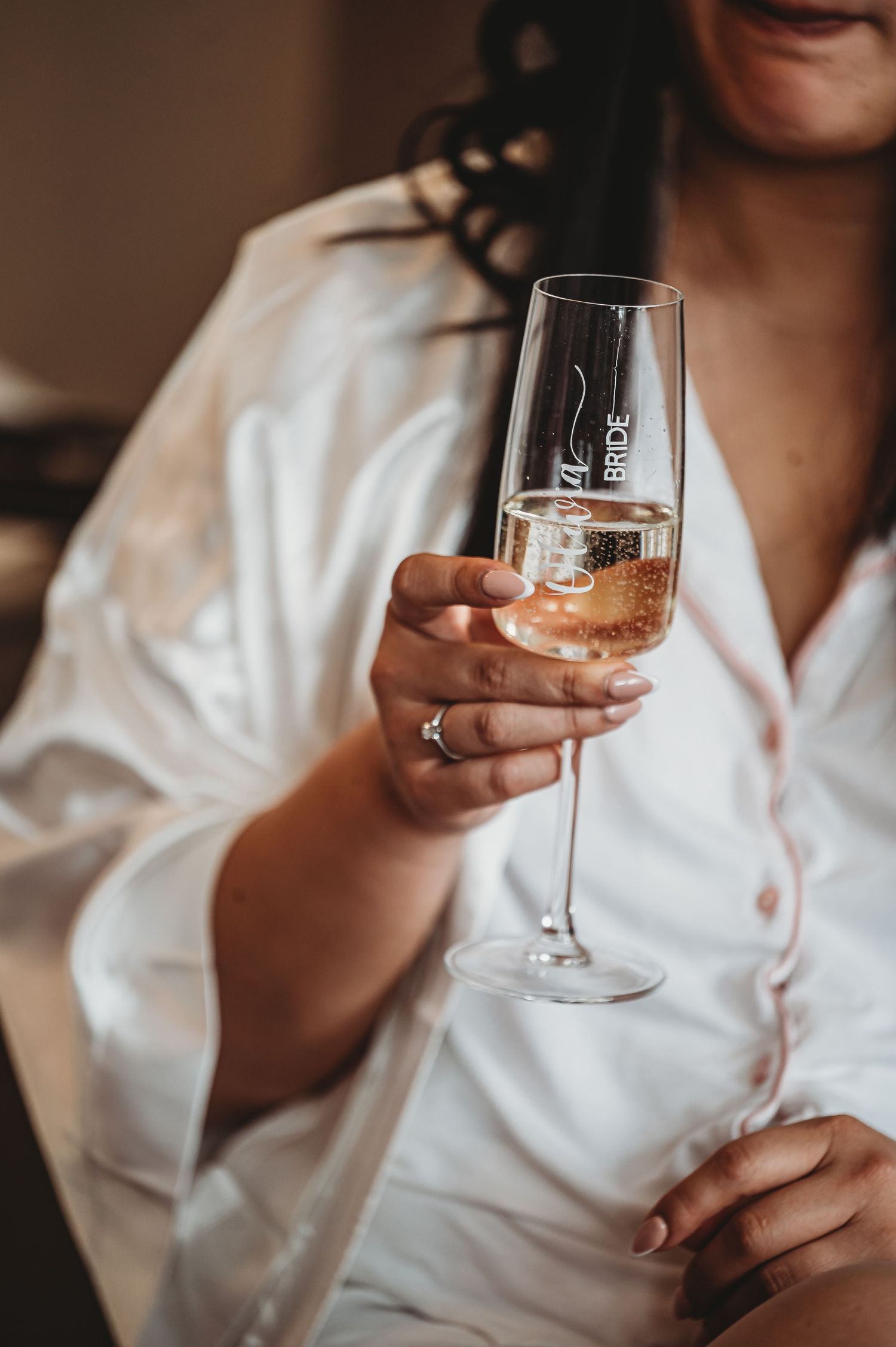 A hand wearing a ring holds an elegant champagne flute filled with sparkling rosé wine against a white shirt.