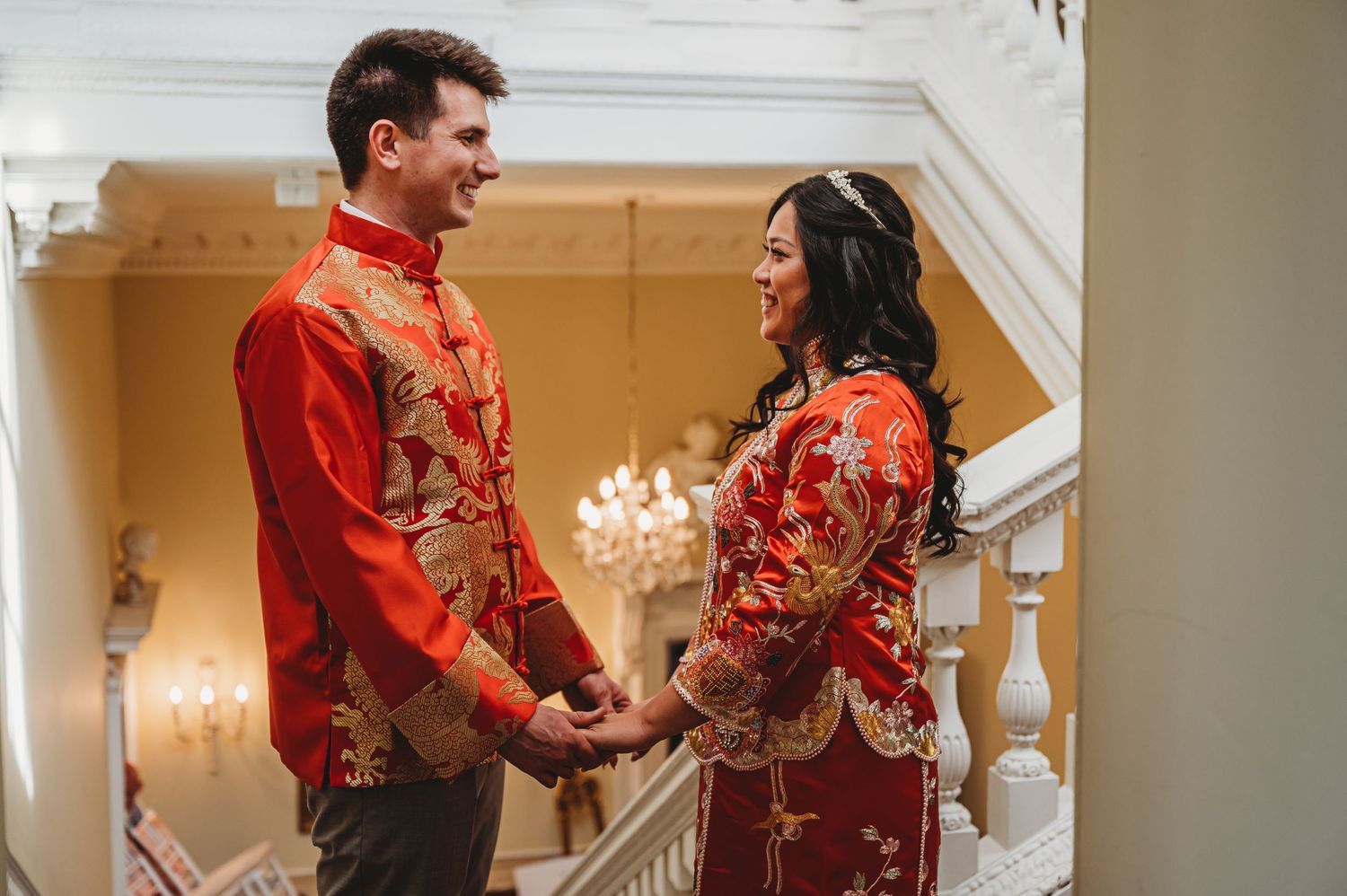 Couple in matching red and gold traditional outfits share intimate moment on elegant staircase.