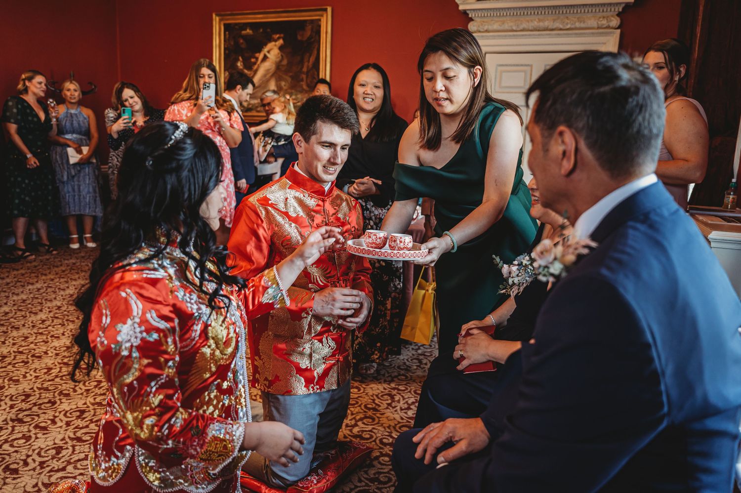 A couple in traditional Asian red floral attire toast drinks with wedding guests during a celebration in an elegant room.
