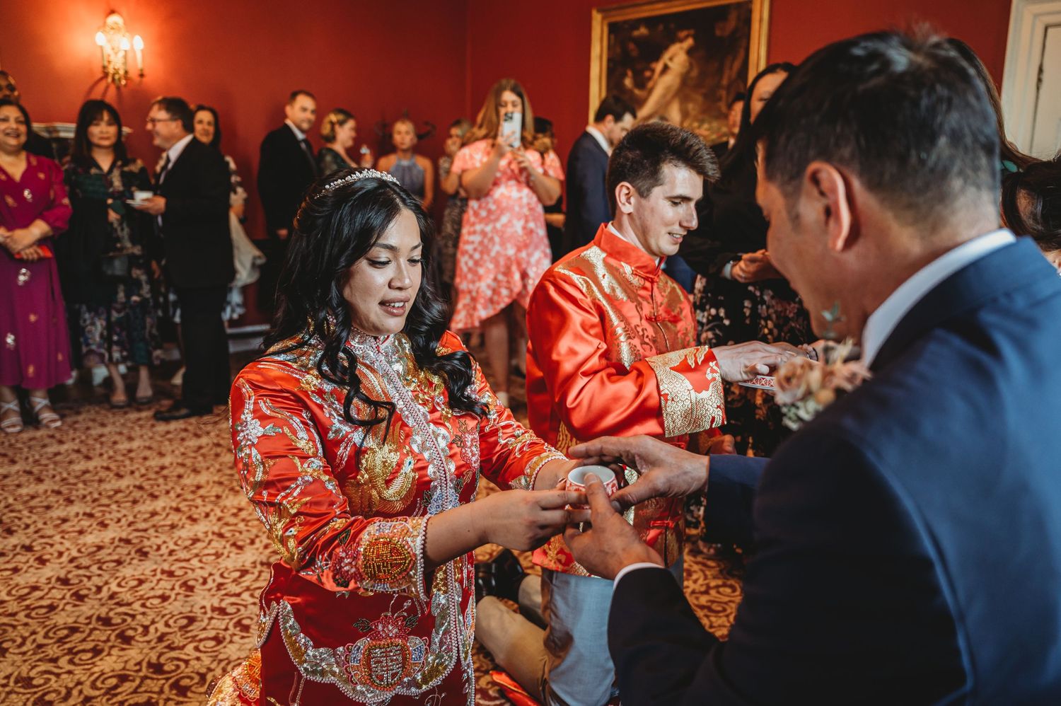 Traditional tea ceremony in red and gold outfits takes place during wedding celebration on ornate carpeted floor.