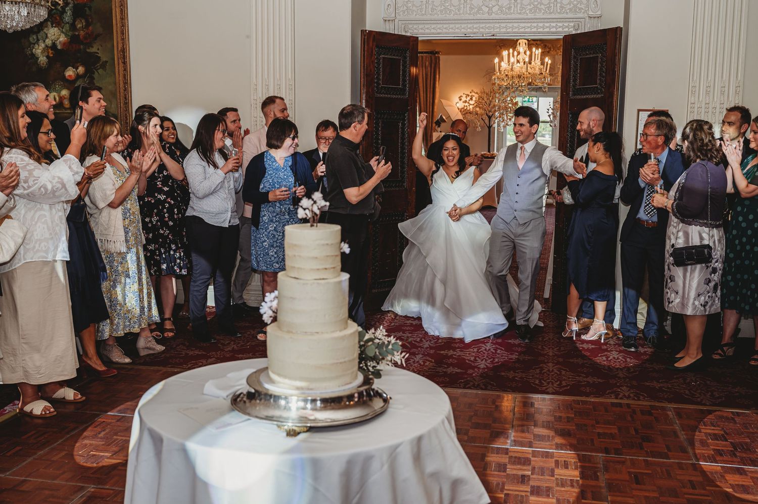 Wedding guests gather around an elegant tiered white wedding cake in an ornate ballroom with chandelier.