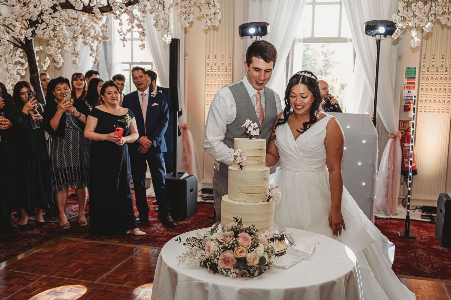 A couple stands next to their wedding cake during their reception celebration in an ornate venue.