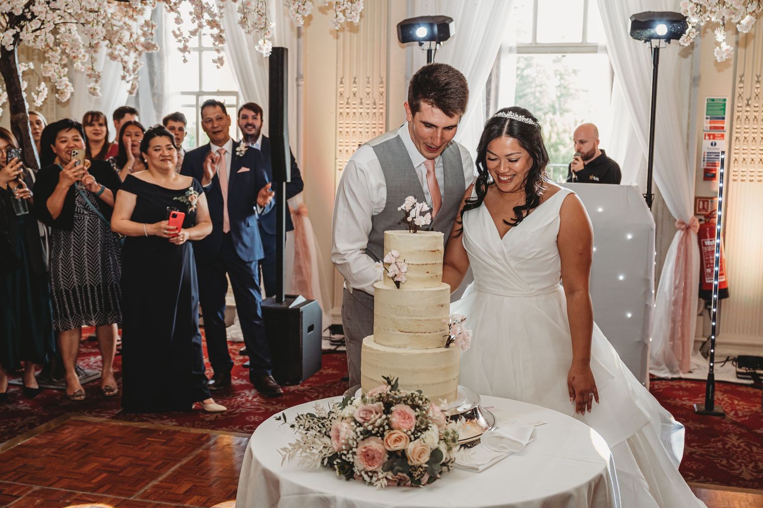 Happy couple cutting their elegant three-tier white wedding cake adorned with pink flowers during reception ceremony.