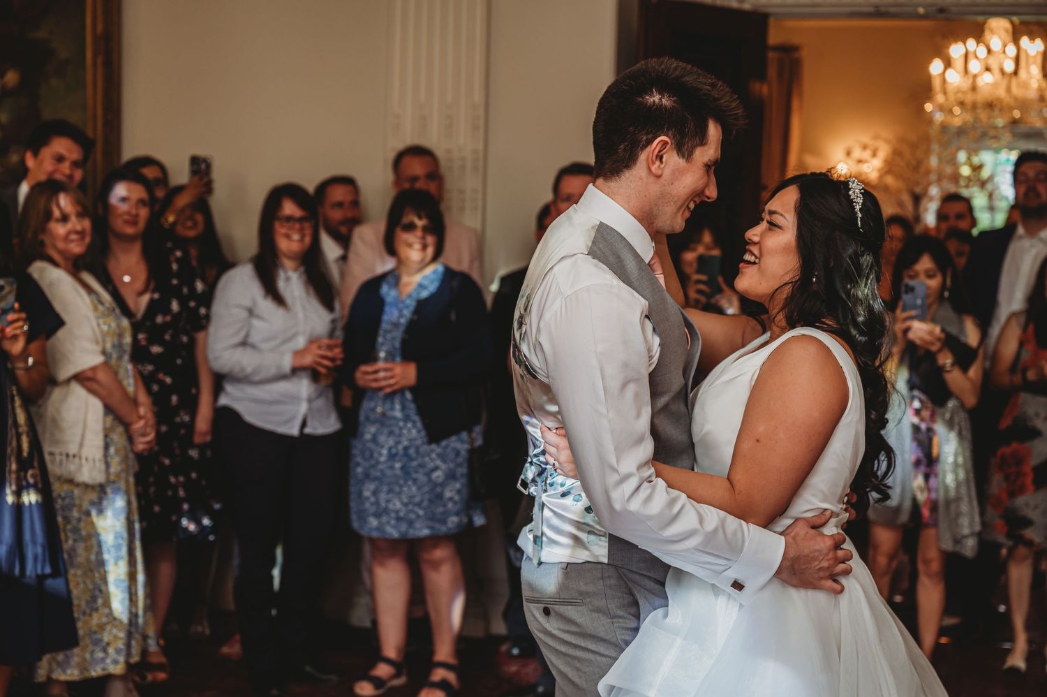 A couple shares their first dance surrounded by wedding guests in an elegant ballroom with chandeliers.