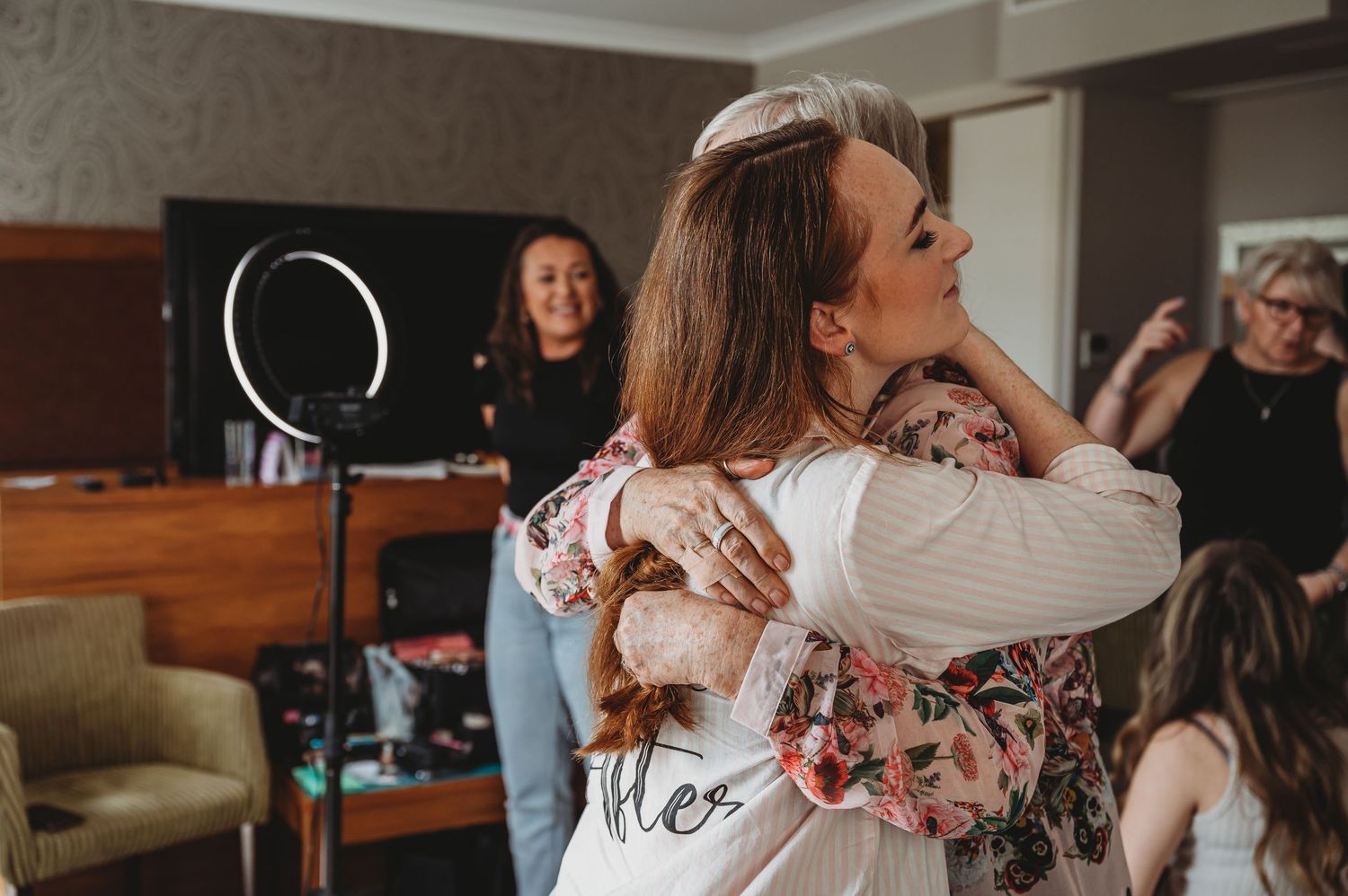 People embracing in an emotional moment during an indoor gathering while others look on in the background with a ring light nearby.
