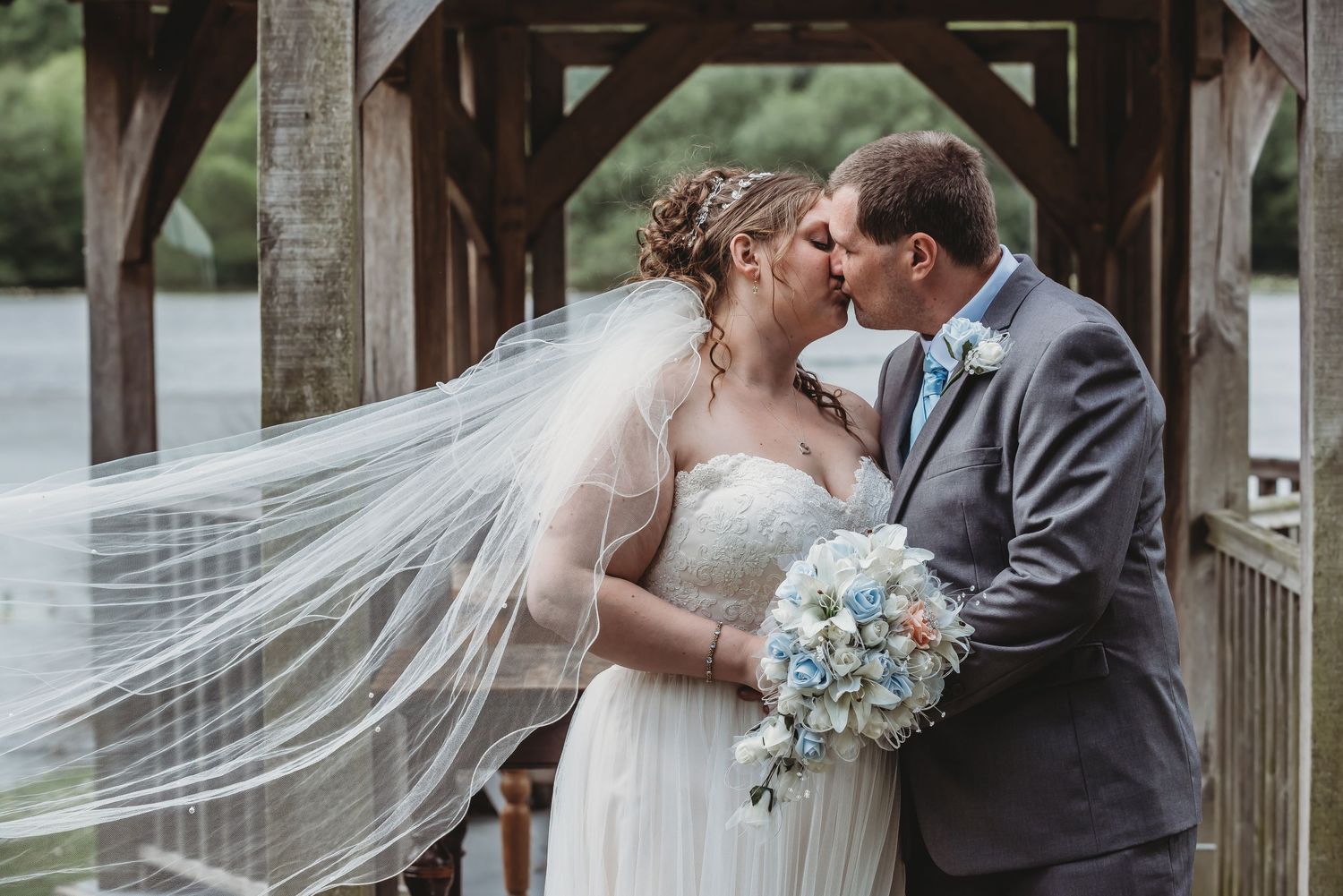 A romantic wedding couple embraces on a wooden bridge with a flowing veil and blue bouquet.