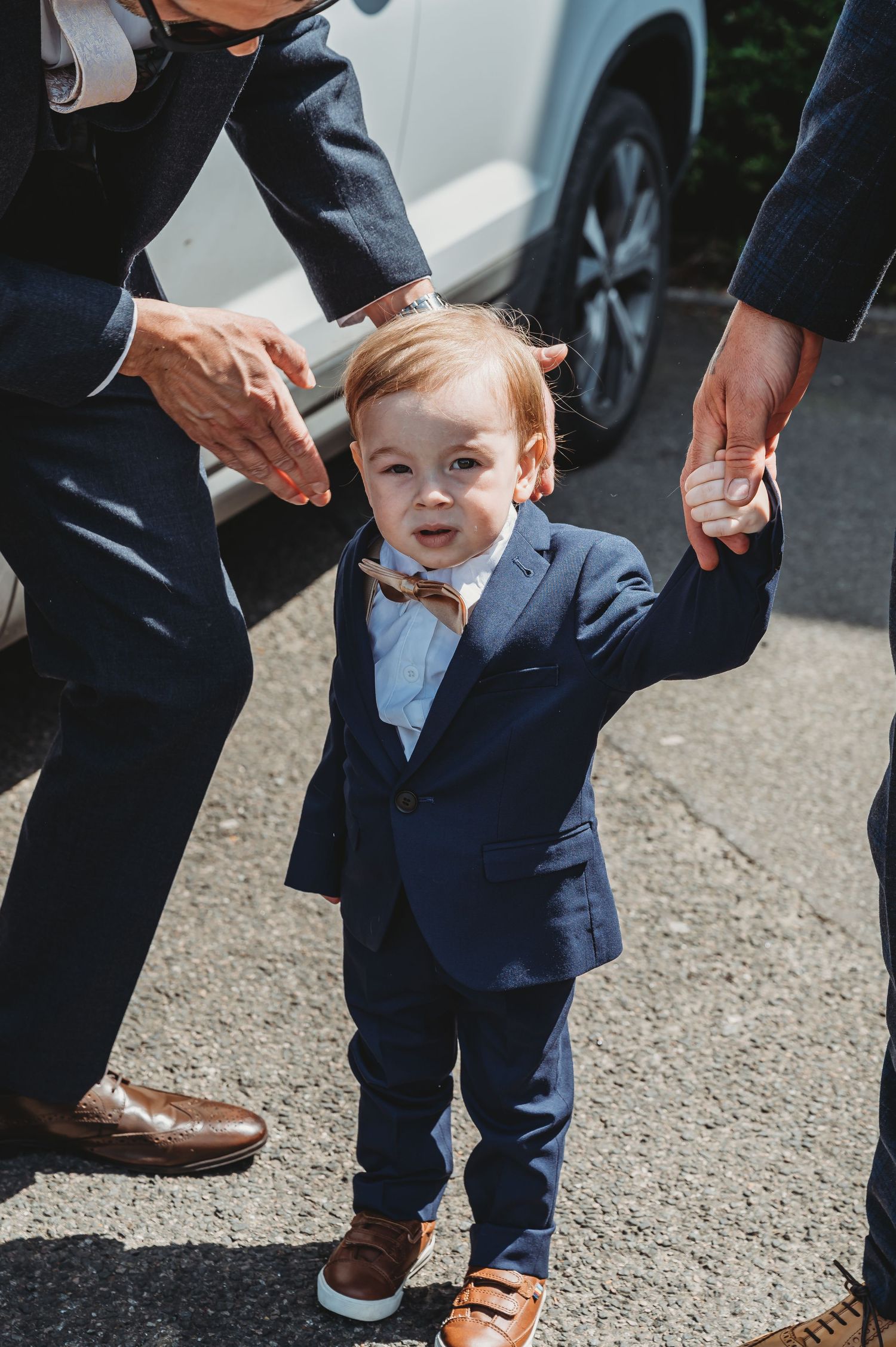 A small child in a navy suit and bow tie holds hands with adults while standing outside near a car.