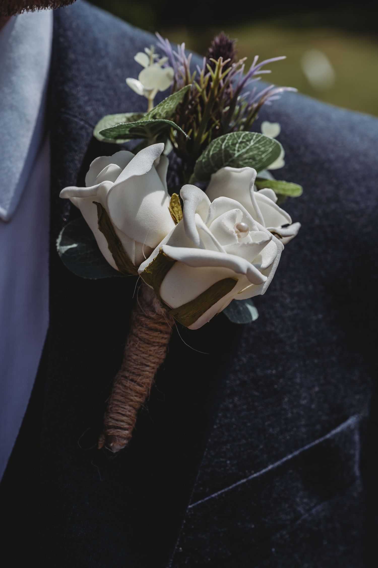 A white rose and purple flower boutonniere wrapped in twine against a dark suit lapel for a formal occasion.