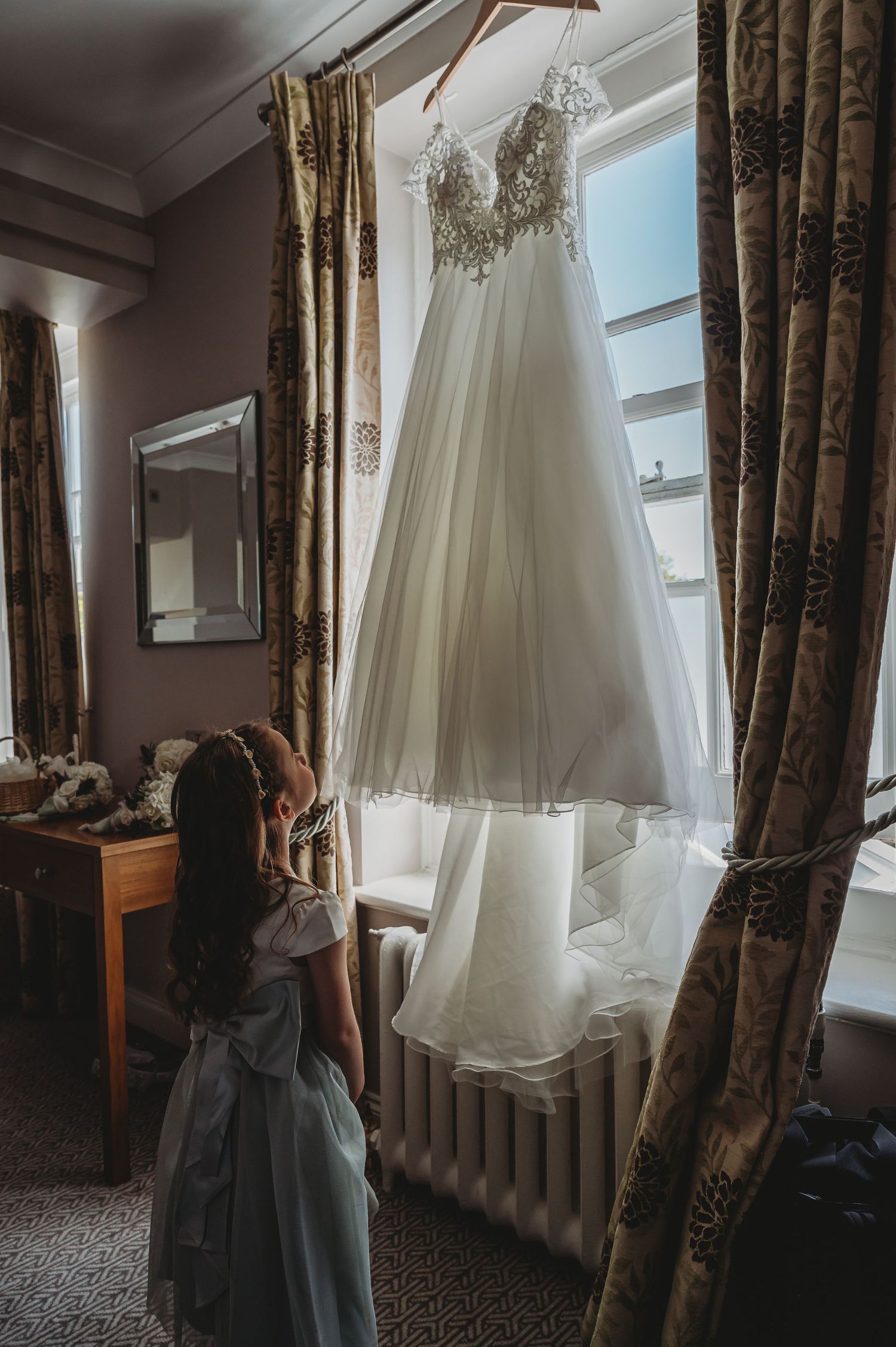 A white lace wedding dress hangs by a window with elegant curtains while someone in gray looks up at it.