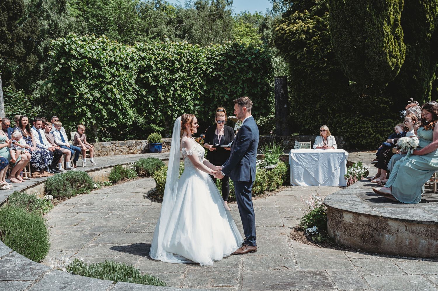 An outdoor garden wedding ceremony with guests seated around a stone patio surrounded by greenery.
