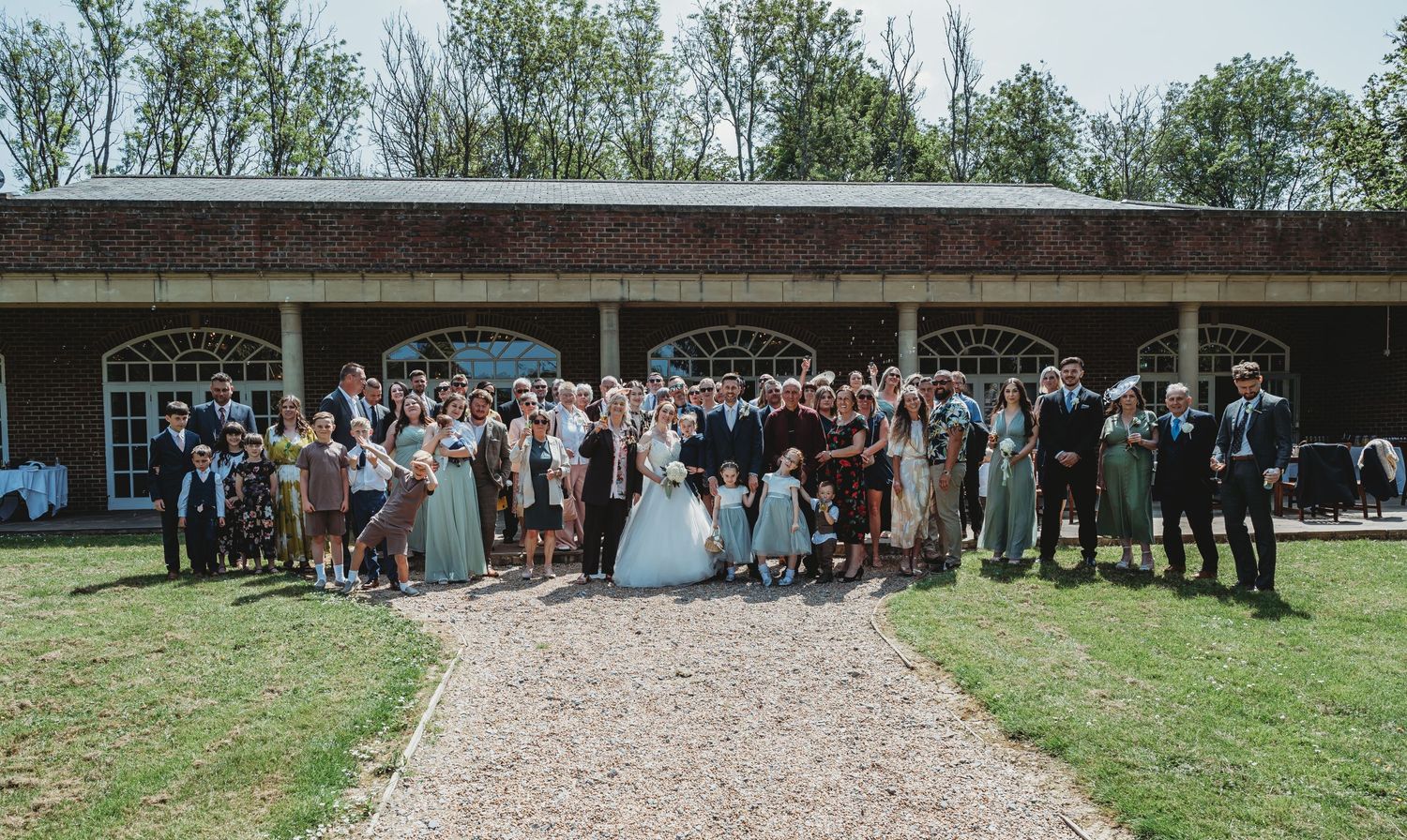 Large outdoor wedding party gathered in front of rustic venue building with gravel path and green lawn.
