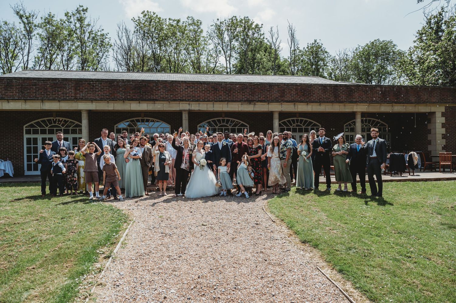 Large group of wedding attendees gathered outside rustic venue building on sunny day with gravel path and green lawn.