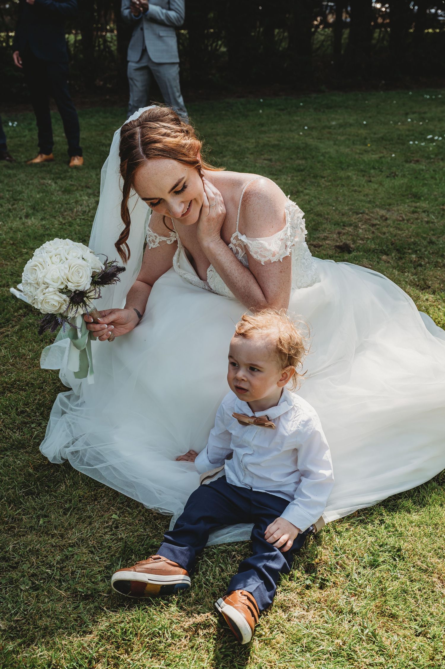 A bride in a white lace wedding dress sits on grass beside a small child wearing a white shirt and bow tie.