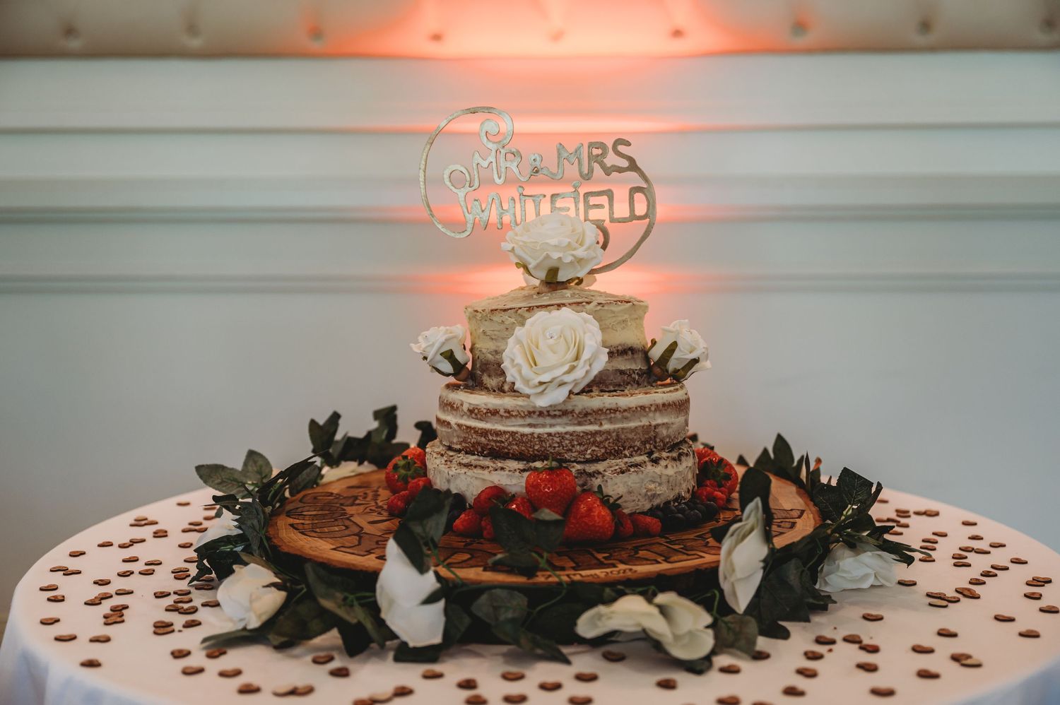 A rustic naked wedding cake decorated with strawberries and greenery, topped with an illuminated neon sign.