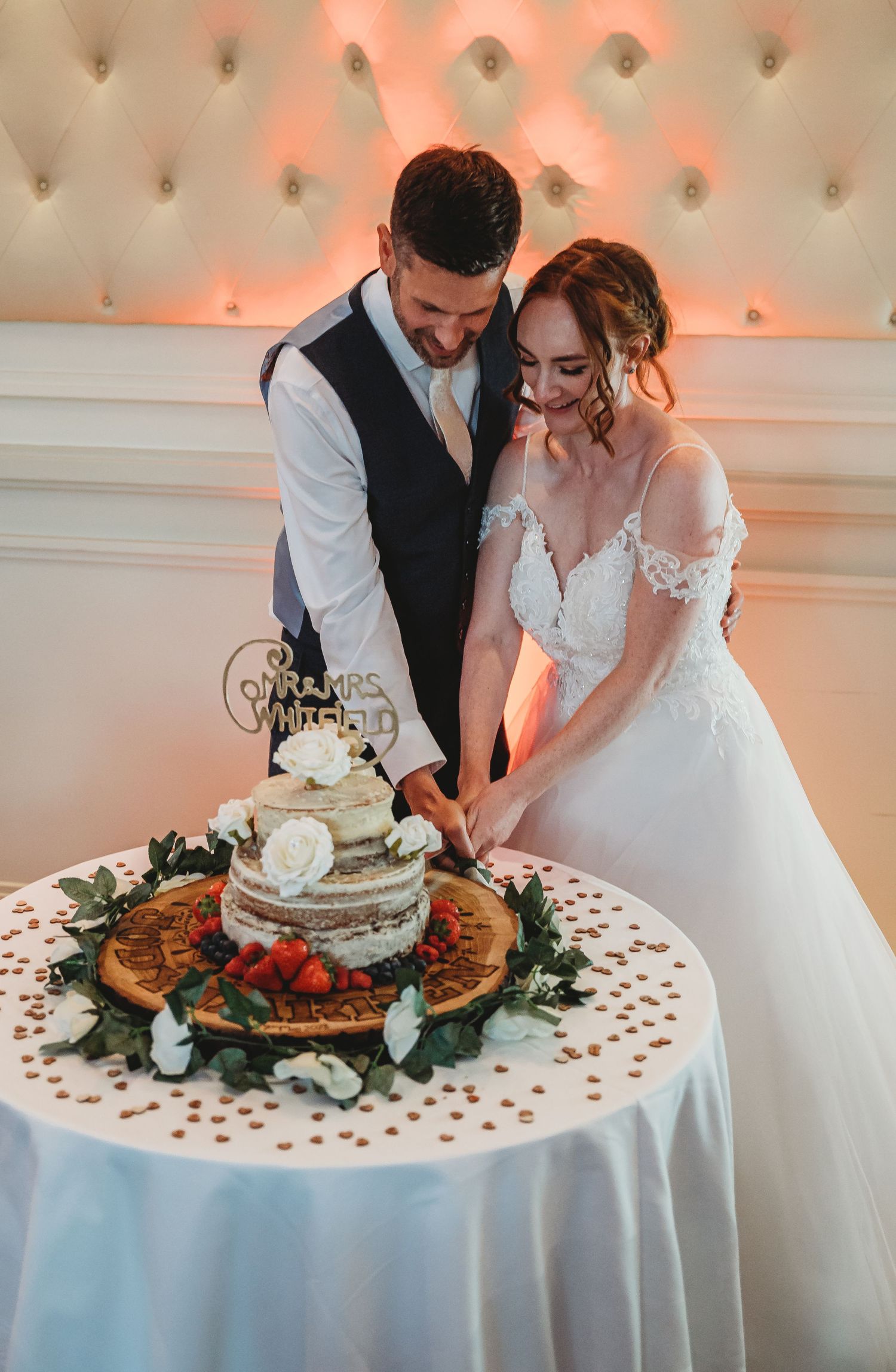 A couple in wedding attire share an intimate moment while cutting their rustic wedding cake decorated with berries and greenery.