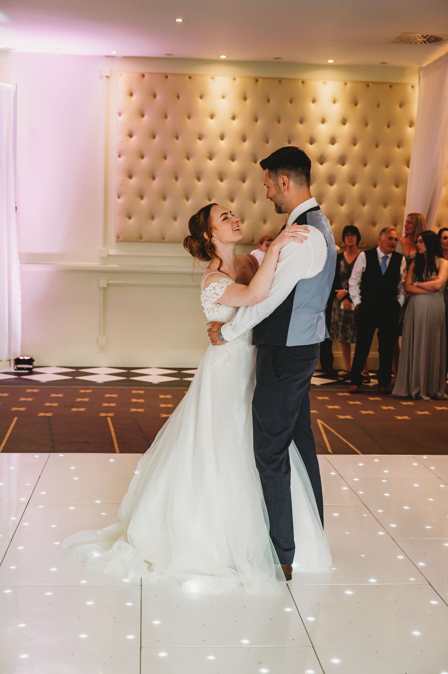 A couple shares their first dance on a lit-up white dance floor with a tufted champagne-colored wall backdrop.