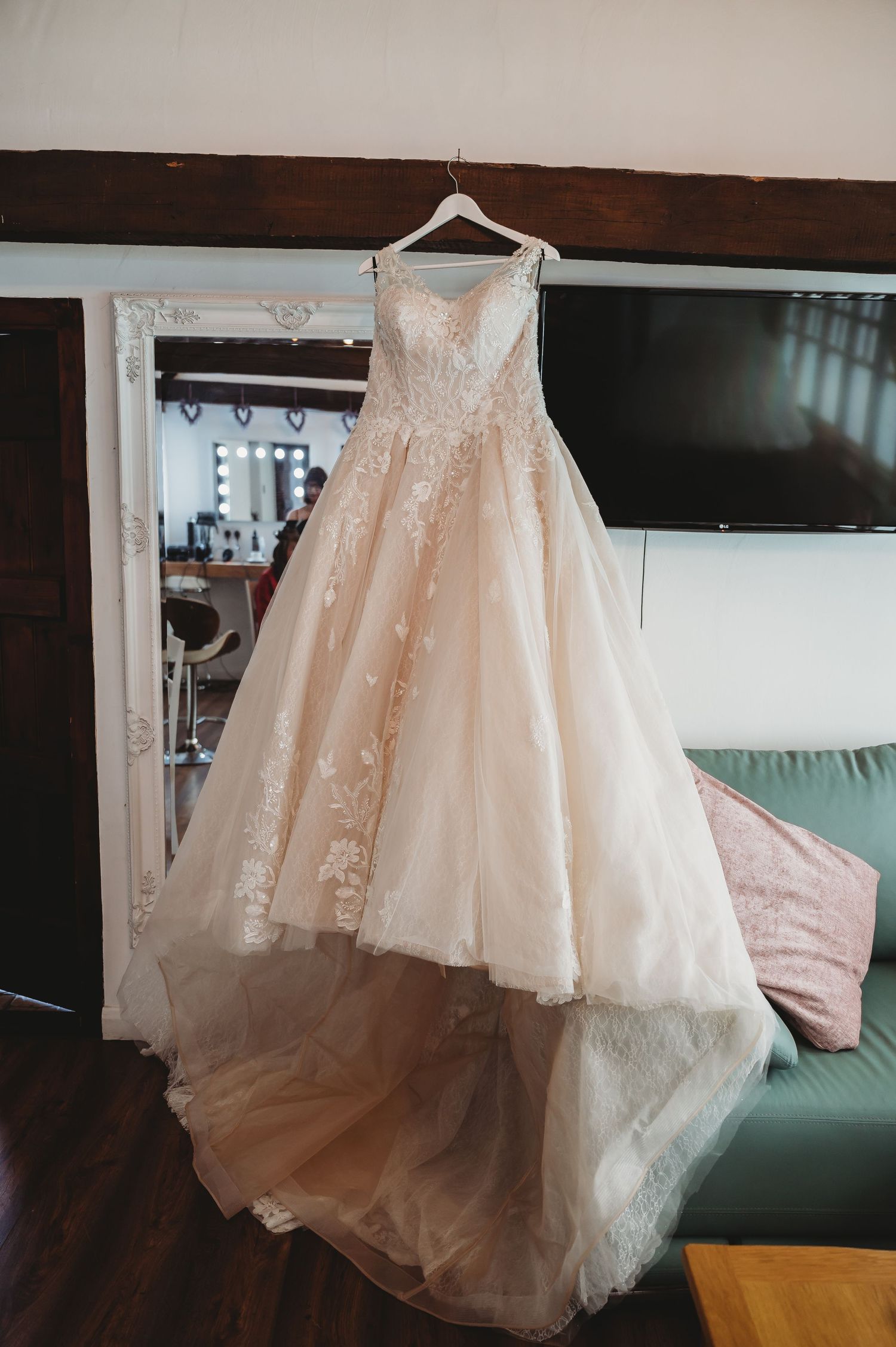 A delicate blush-colored wedding gown with lace detailing hangs on display against a wall with a TV in the background.