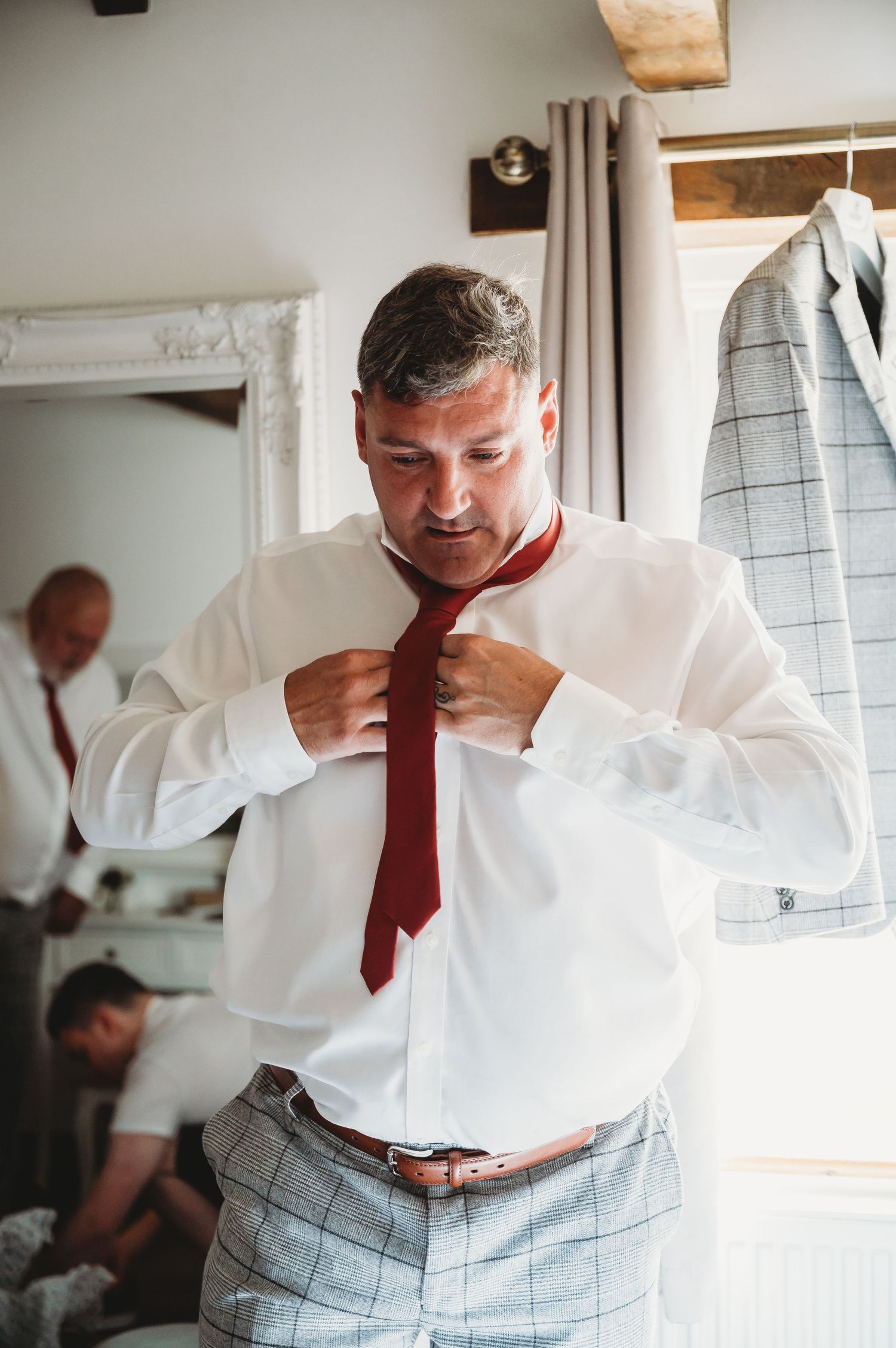 Someone adjusts a red tie while getting dressed in formal attire with grey plaid pants and white dress shirt.