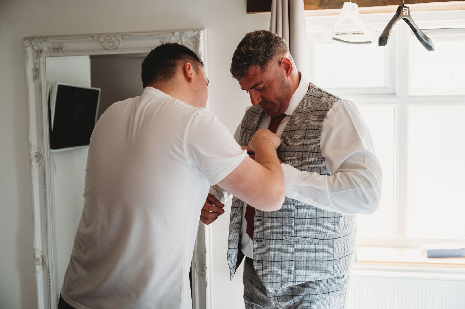 Two people getting ready for a formal event as one helps adjust the other's gray plaid vest in a bright room.