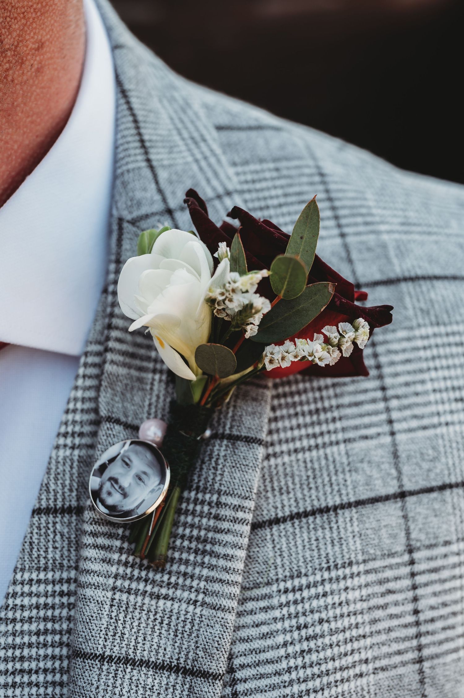 A wedding boutonniere with white rose and dark burgundy accents pinned to a gray plaid suit jacket.