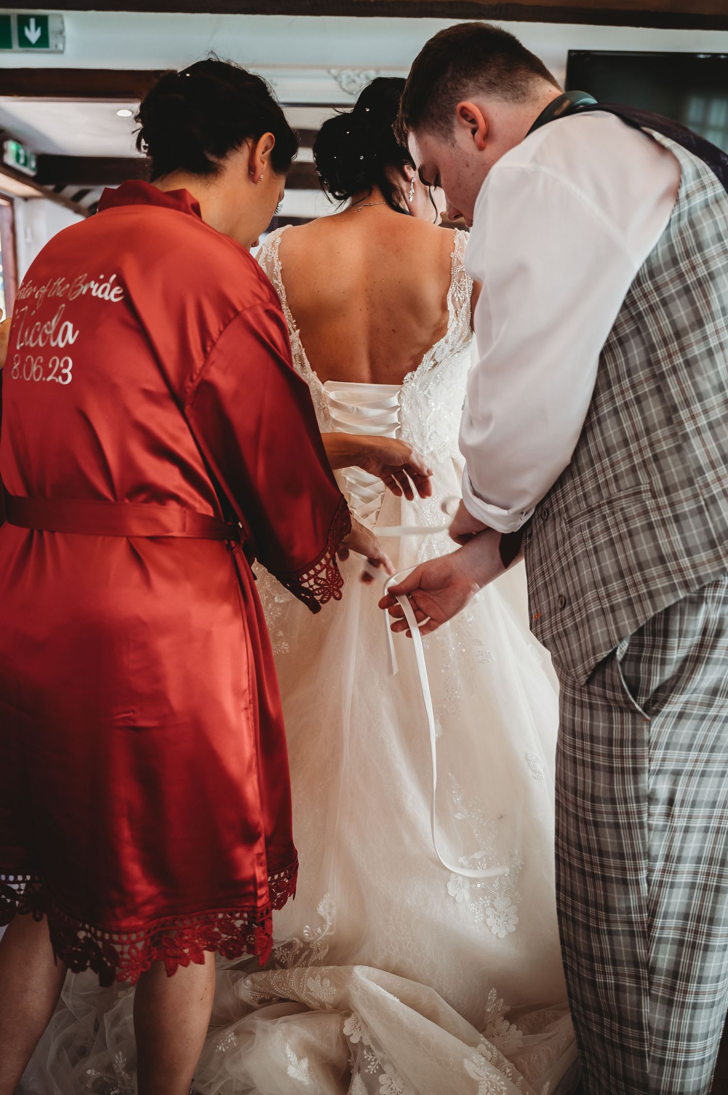 Two people help adjust a white lace wedding dress during wedding preparations.