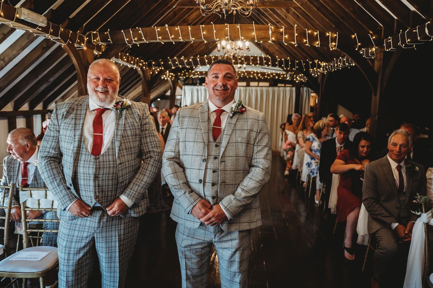 Two wedding attendants in matching light gray plaid suits stand in a rustic barn venue decorated with string lights.