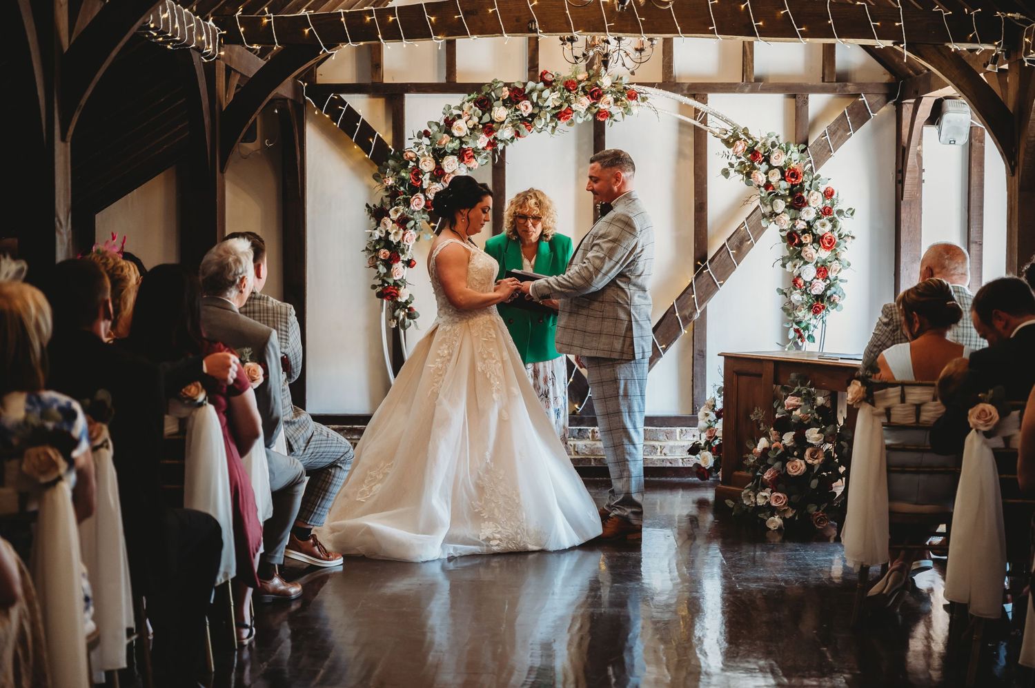A couple exchanges vows under a floral arch during their rustic barn wedding ceremony with guests watching from wooden pews.