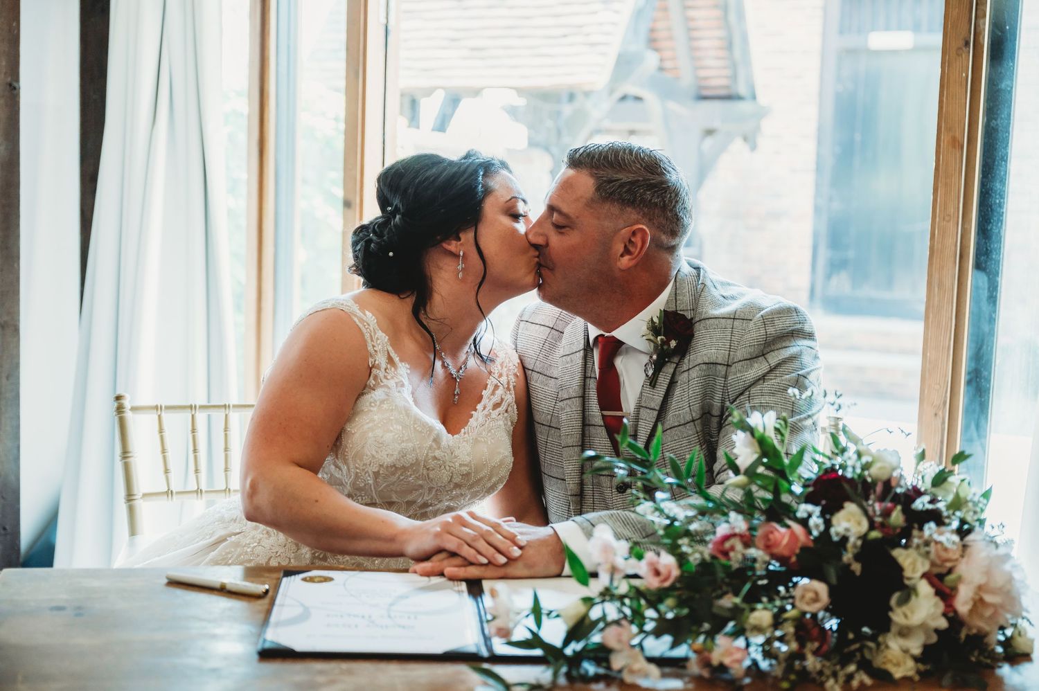 Two people share a tender kiss while seated at a table with a floral bouquet and documents.