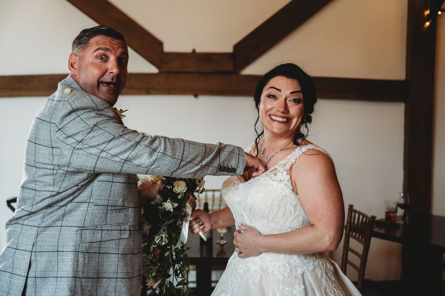 A couple shares a lighthearted moment at a rustic wedding venue with exposed wooden beams and floral decor.