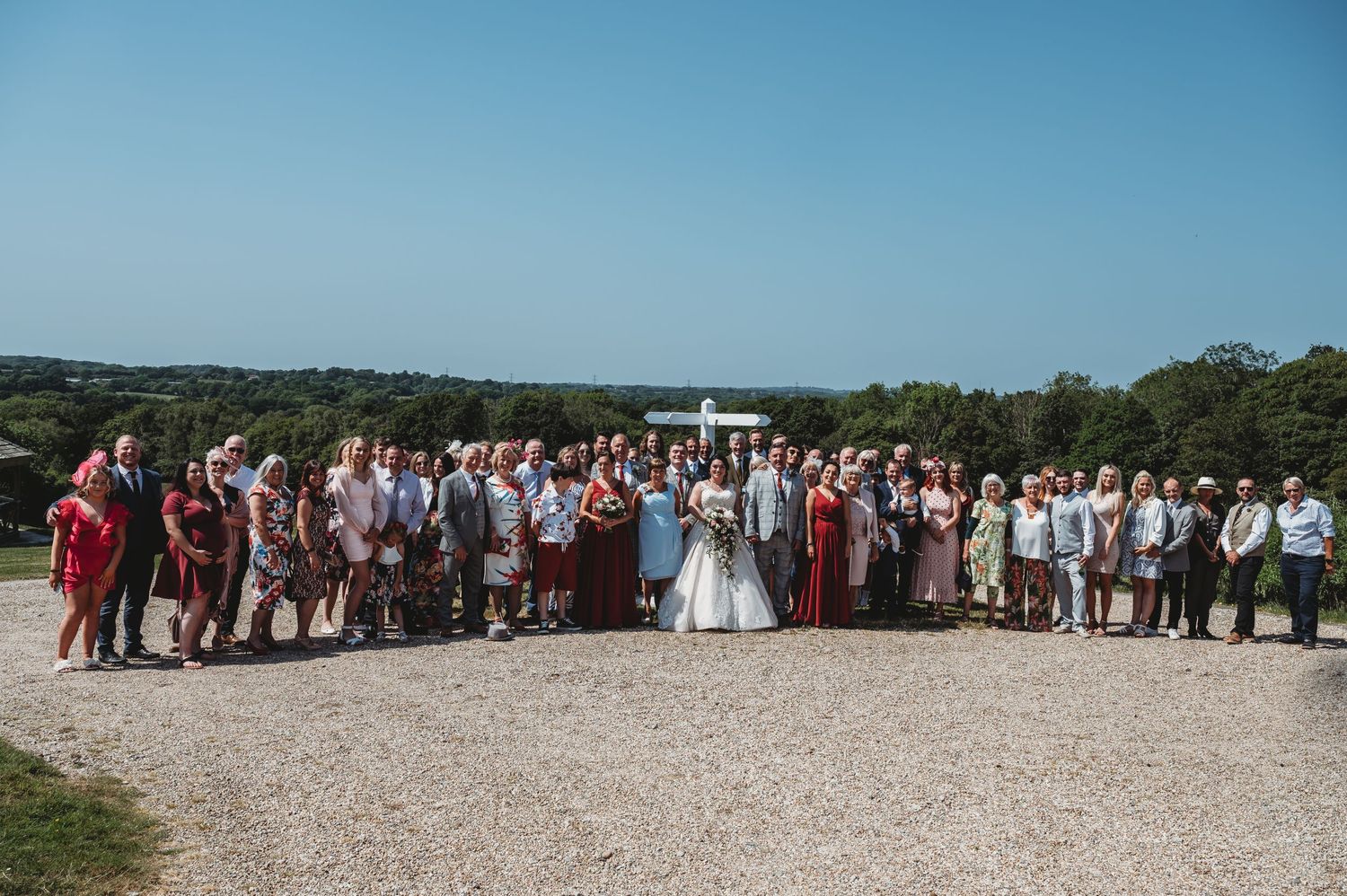 Large wedding party gathered on a gravel path for outdoor group photo with blue sky and green trees in background.