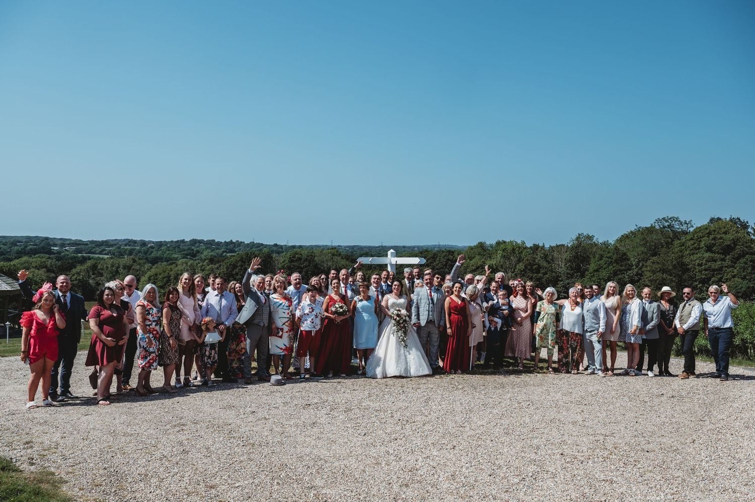 Wedding party and guests gathered on a gravel path at a scenic outdoor venue on a bright sunny day.