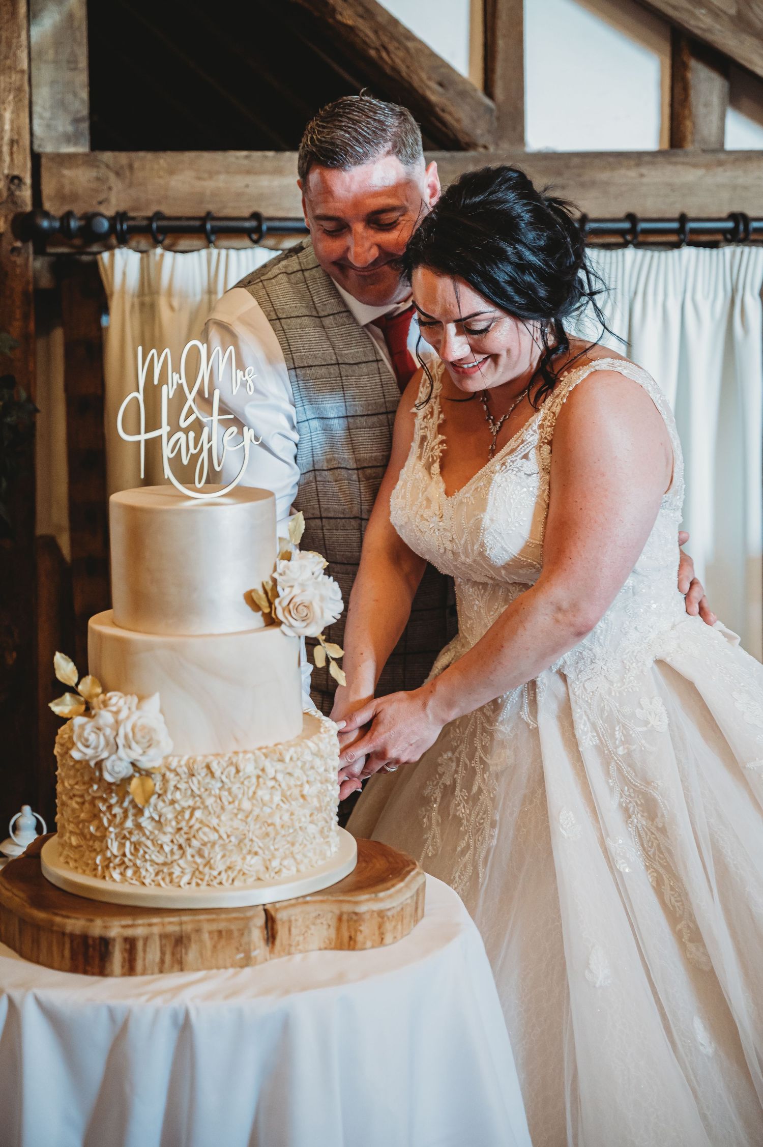 A couple cuts their elegant three-tier white wedding cake decorated with lace details and floral accents on a rustic wood stand.