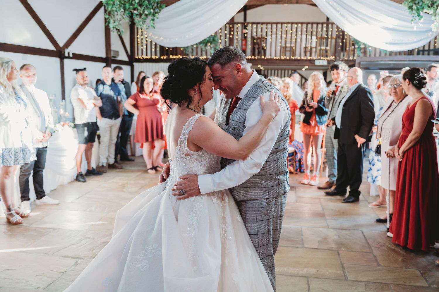 Couple shares romantic first dance together in rustic barn venue surrounded by wedding guests in coral dresses.
