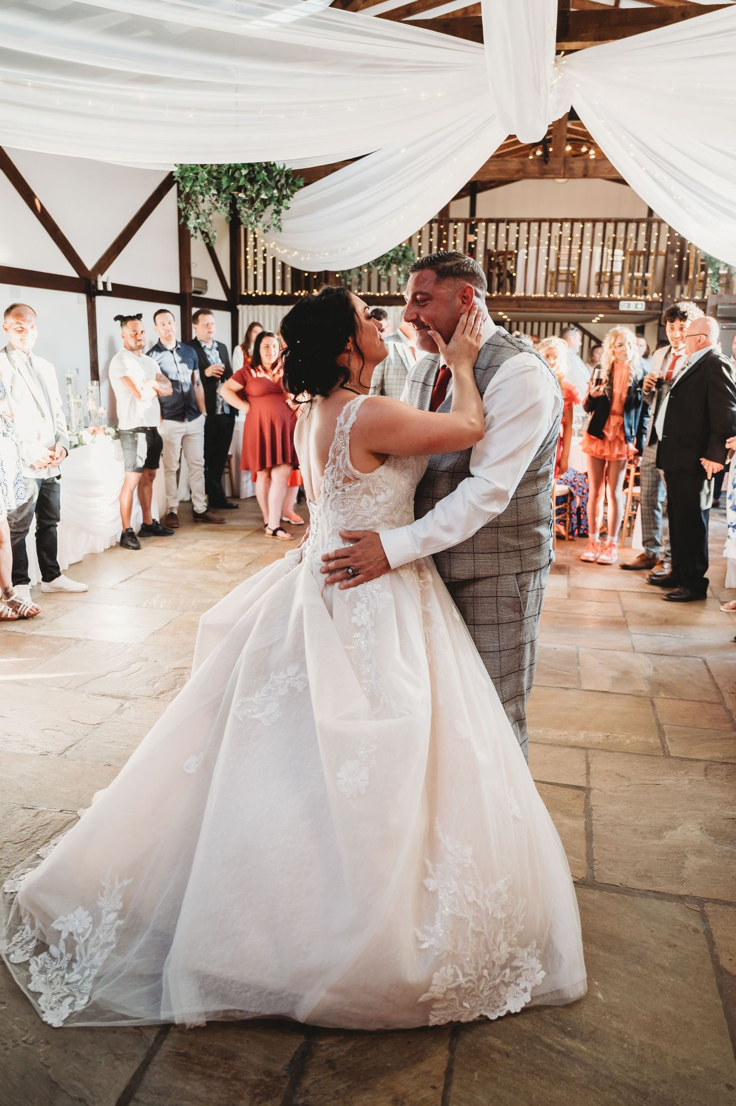 A romantic wedding couple shares their first dance under white draping and string lights in a rustic barn venue.