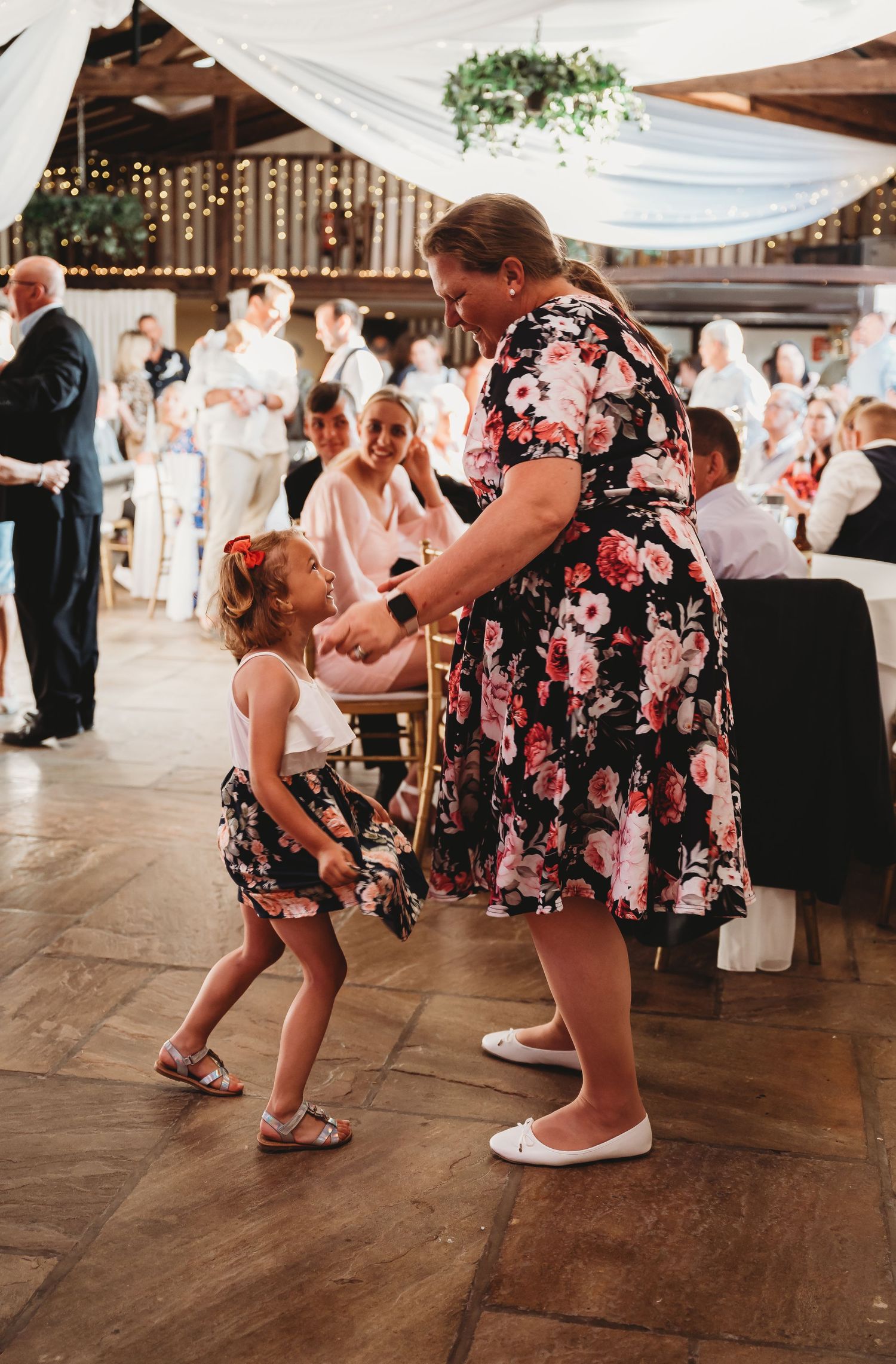 Two people in matching floral dresses dance together on a wooden floor at a wedding reception decorated with string lights.