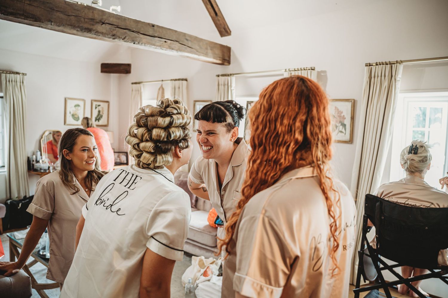 Three people in beige and white outfits gather indoors near a stone fireplace in a rustic room with wooden beams.