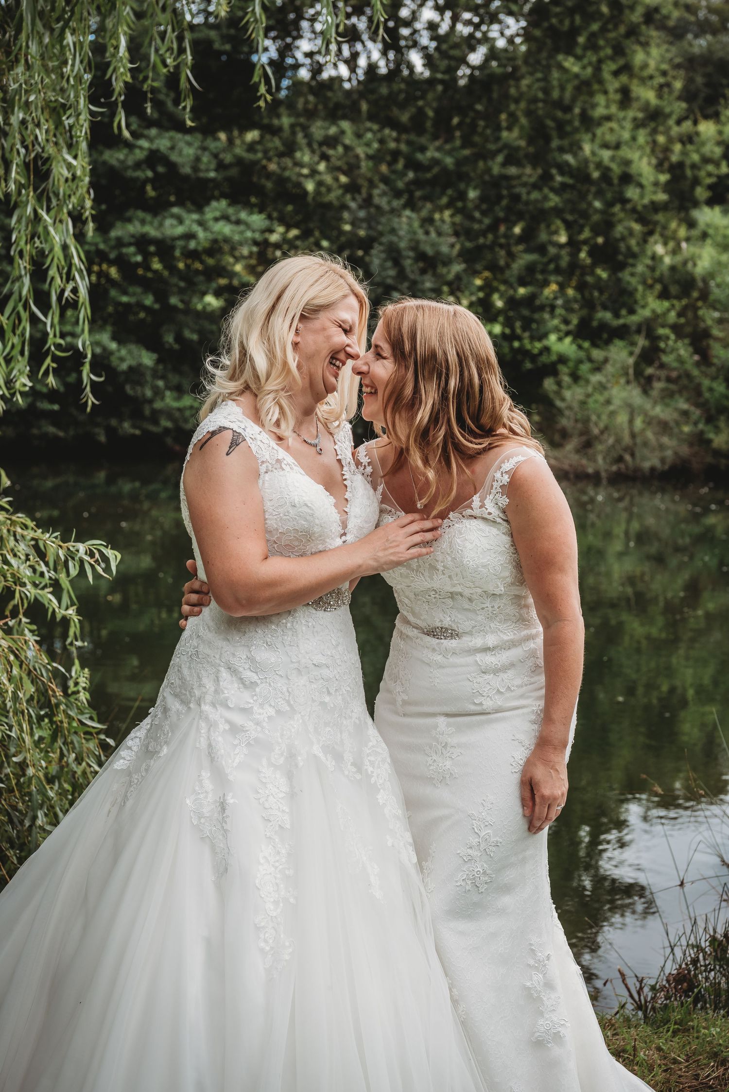Two brides in white wedding dresses share a joyful moment together by a pond under a willow tree.