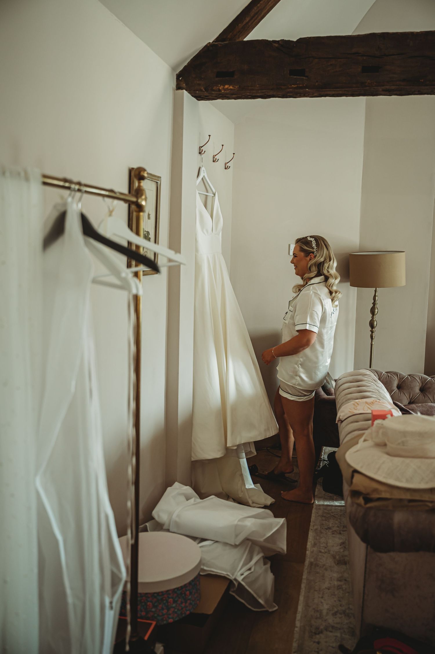 A white wedding dress hangs on display in a rustic room while someone in casual attire stands nearby.