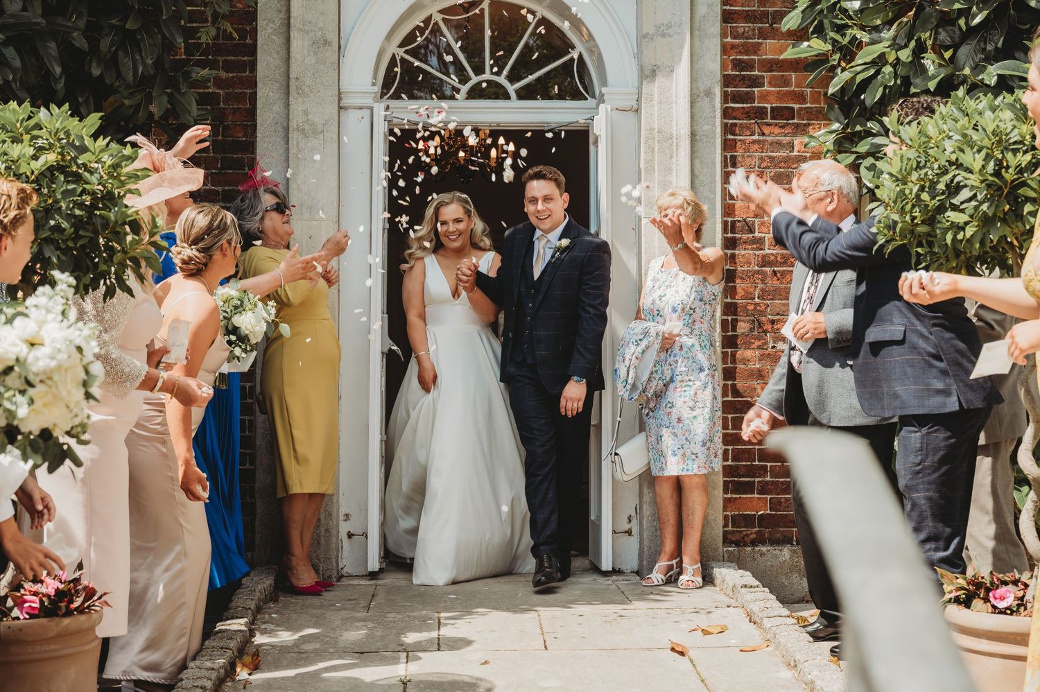 Wedding party celebrates with confetti as happy couple exits historic brick church doorway in joyful ceremony moment.