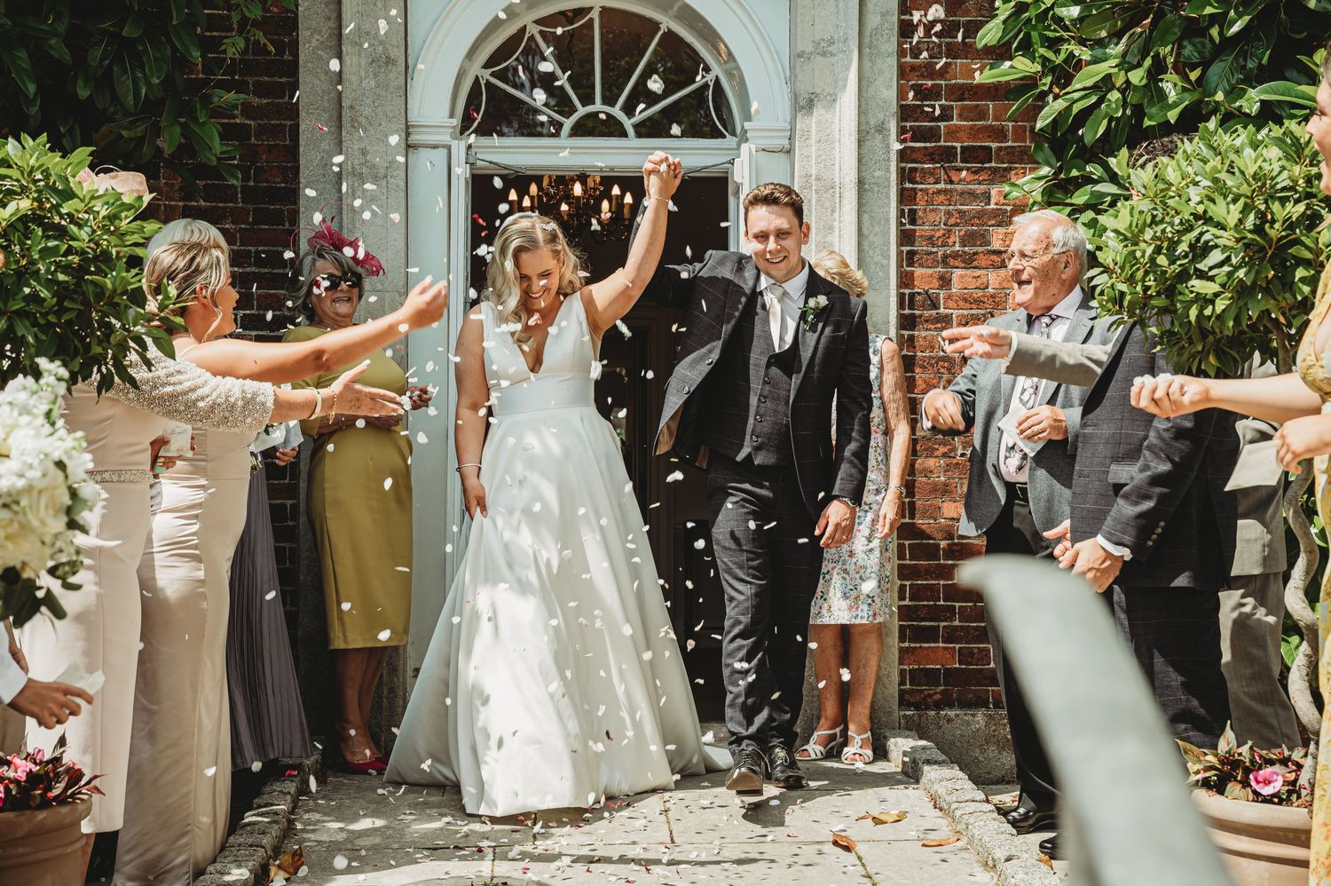 Wedding couple exits ceremony at brick venue with joyful arms raised while guests toss confetti around them.
