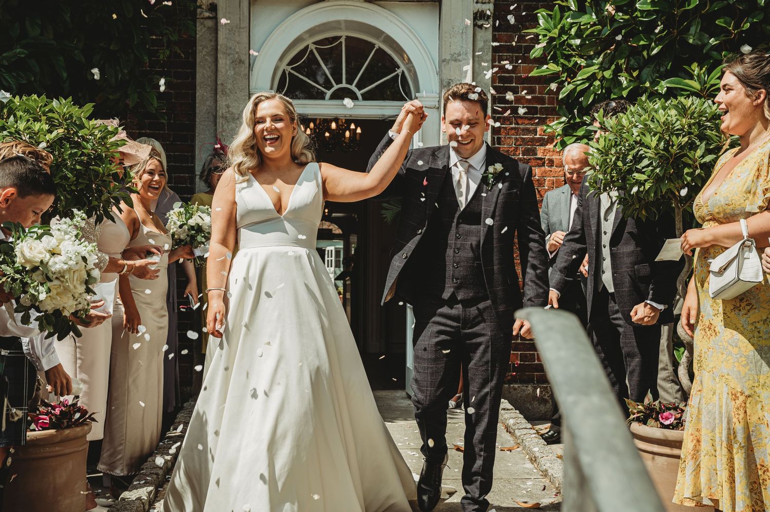 Wedding couple exits historic venue as guests throw confetti during celebratory recessional in garden setting.