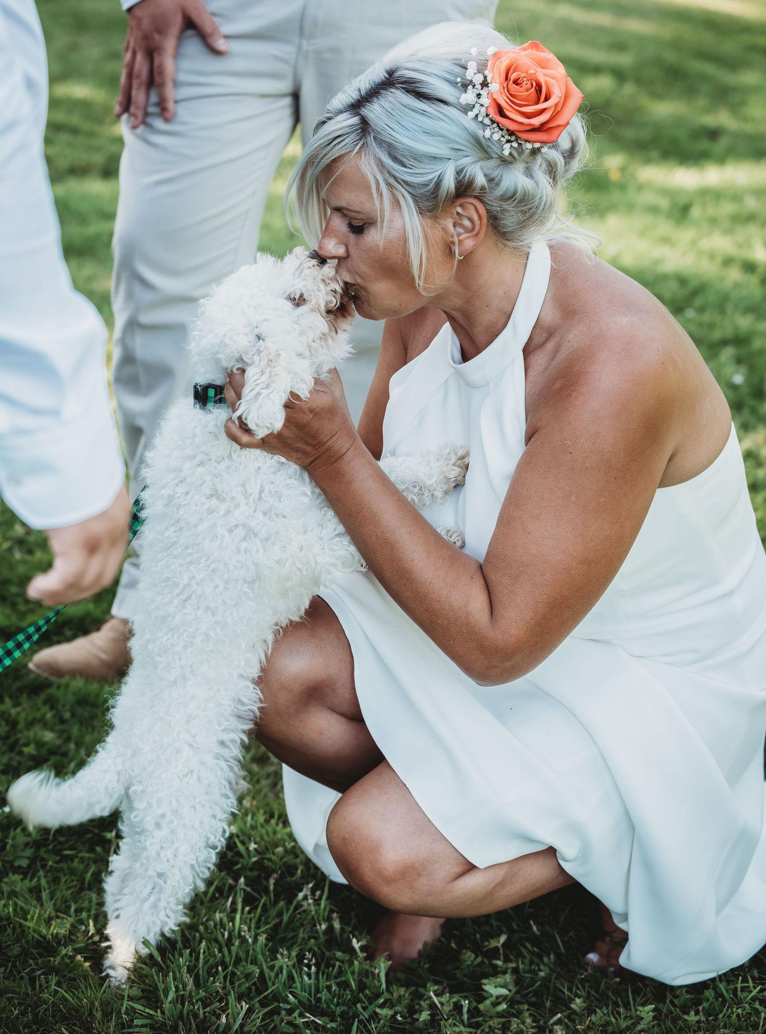 A bride in a white dress embraces and kisses a fluffy white dog on green grass during a wedding celebration.