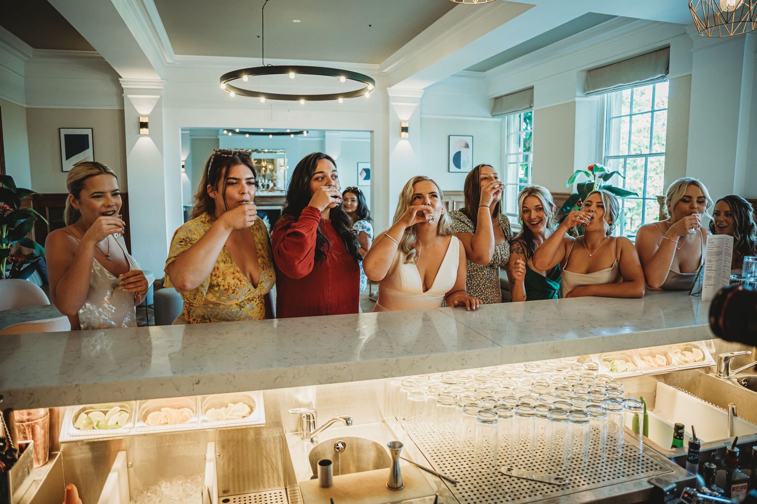 A group of wedding guests laugh together while leaning on an illuminated marble bar counter at a reception.