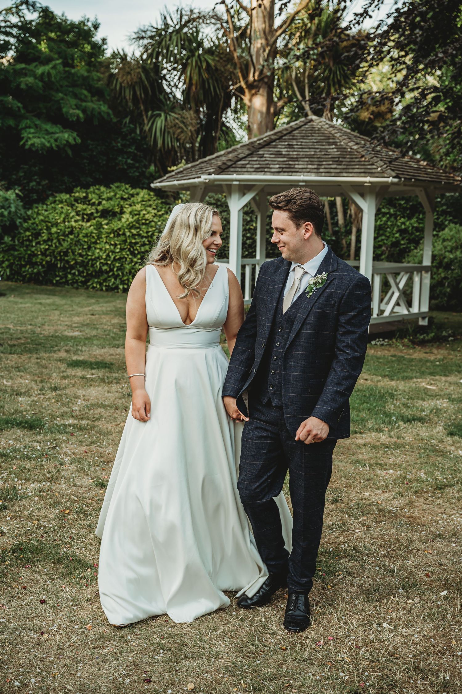 A couple in wedding attire stand together on a lawn near a white wooden gazebo surrounded by lush garden landscaping.