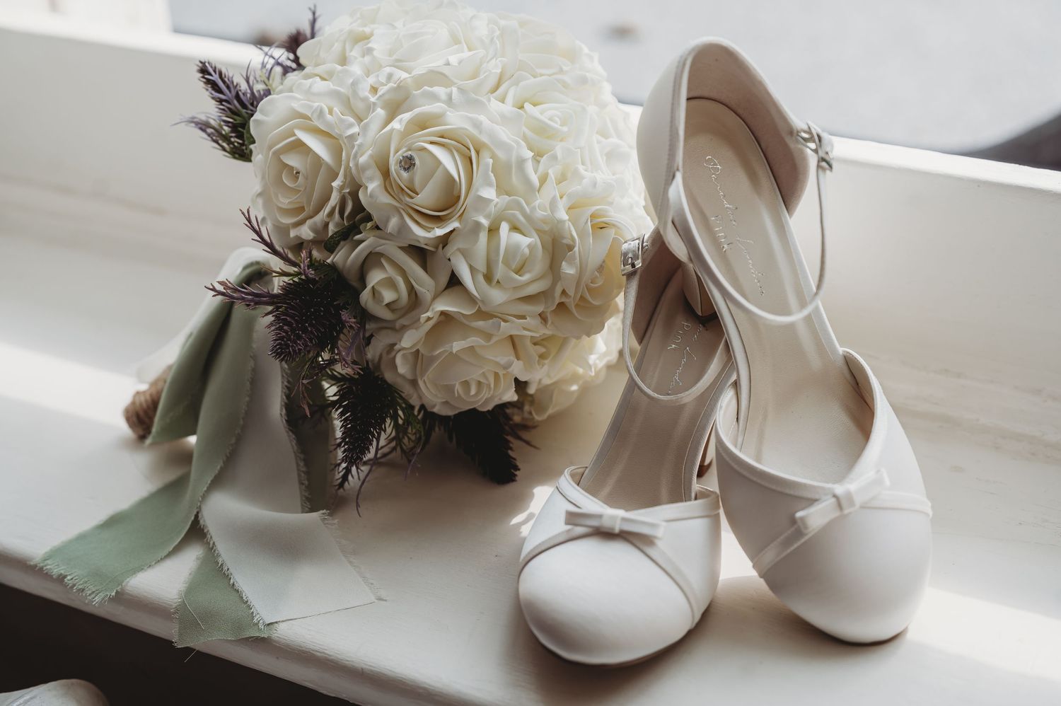 White bridal shoes and a romantic bouquet of white roses with sage green ribbons sit on a windowsill.