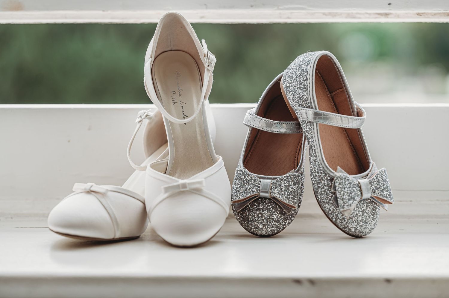 Two pairs of dress shoes, one white satin ballet flat and one silver glitter Mary Jane style, displayed on windowsill.