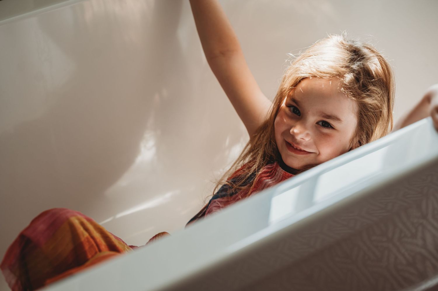 A happy child plays and reaches upward while sitting in a white bathtub.
