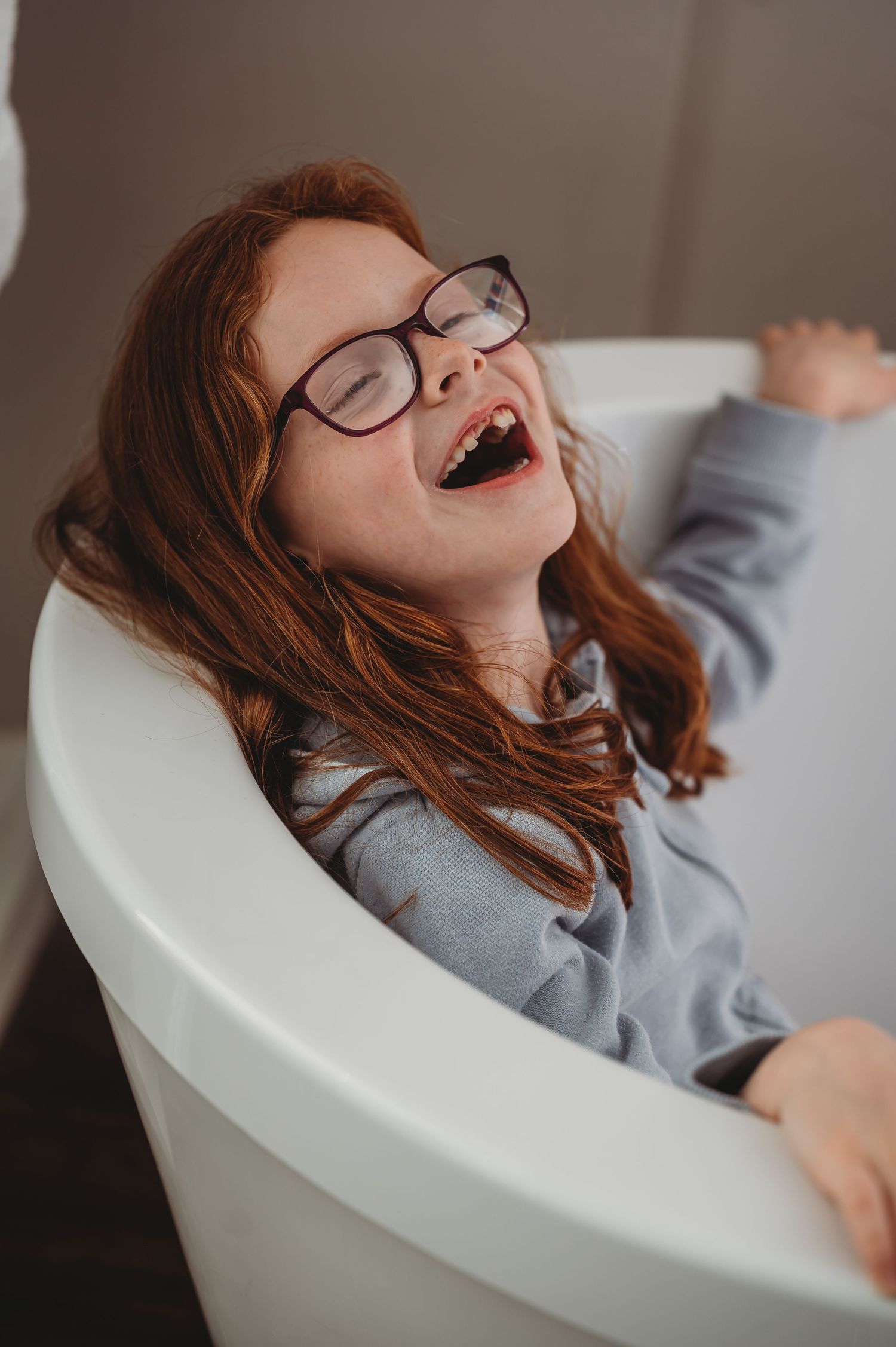 Candid portrait of someone with glasses and red hair laughing joyfully while relaxing in a white chair.