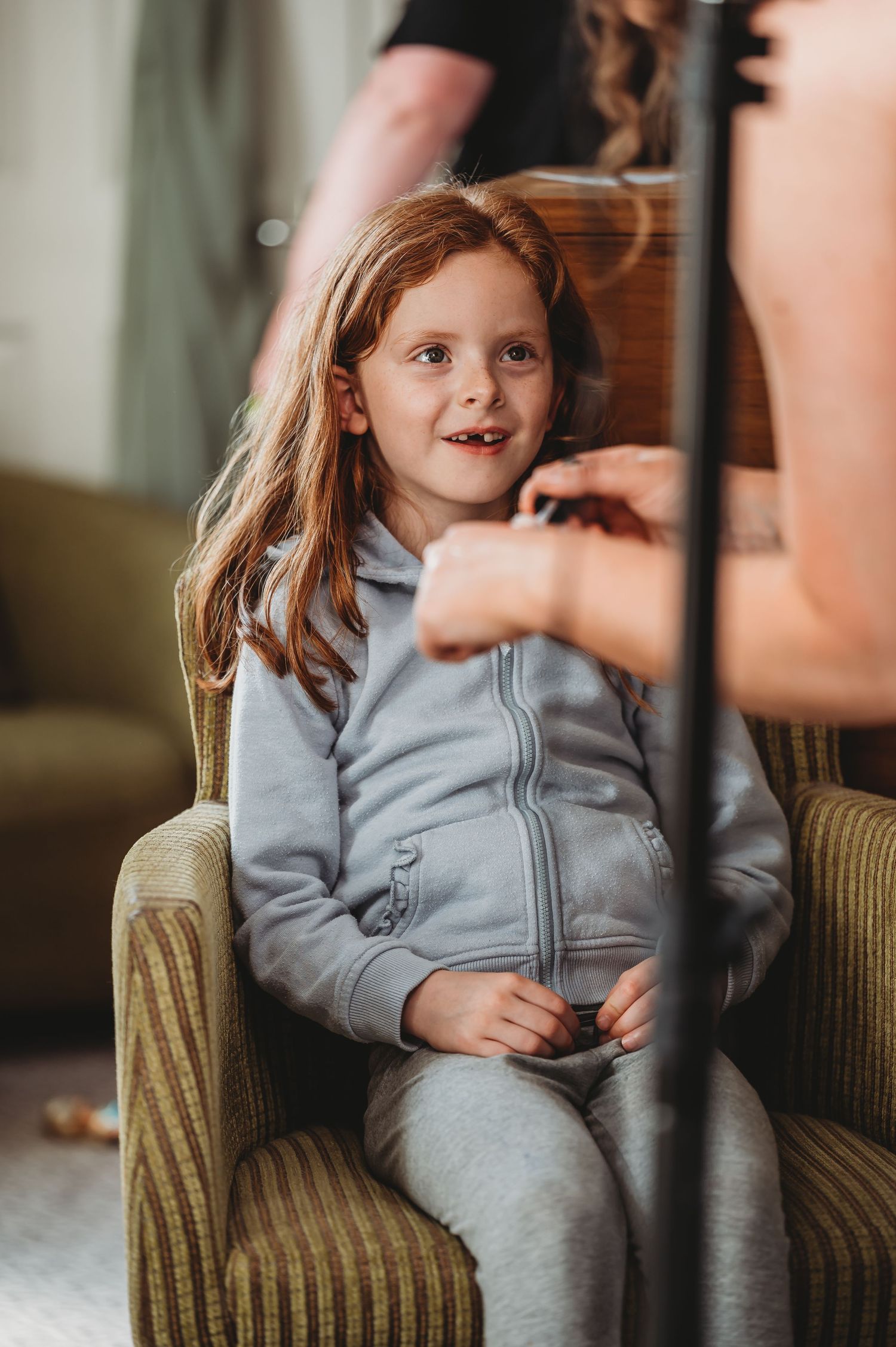 Child in a gray zip-up sweatsuit sits in a striped armchair while getting hair styled.